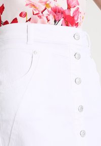 Jupe en denim blanc avec une taille ajustée, fermeture à boutons à l'avant et poches latérales. Présente des finitions en métal argenté et une texture lisse.