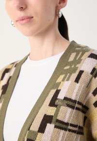 Adīts kardigāns, kas izceļas ar ģeometrisku rakstu olīvzaļā, brūna un melnā krāsā. V-veida izgriezums ar ribu apdari un mazu logotipu priekšpusē.
