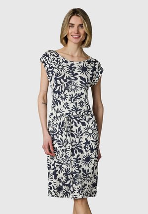 Frau lächelt, trägt ein weißes kurzärmeliges Kleid mit schwarzem abstraktem Blumenmuster und einem Tunnelzug in der Taille, steht vor hellem Hintergrund.