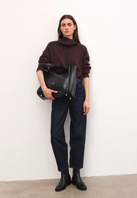 Naine, kes kannab pruunist kõrge kaelusega sviitrit, tumedaid siniseid teksaseid ja musti saapaid, hoiab suurt musta nahast käekotti valge seina vastas.