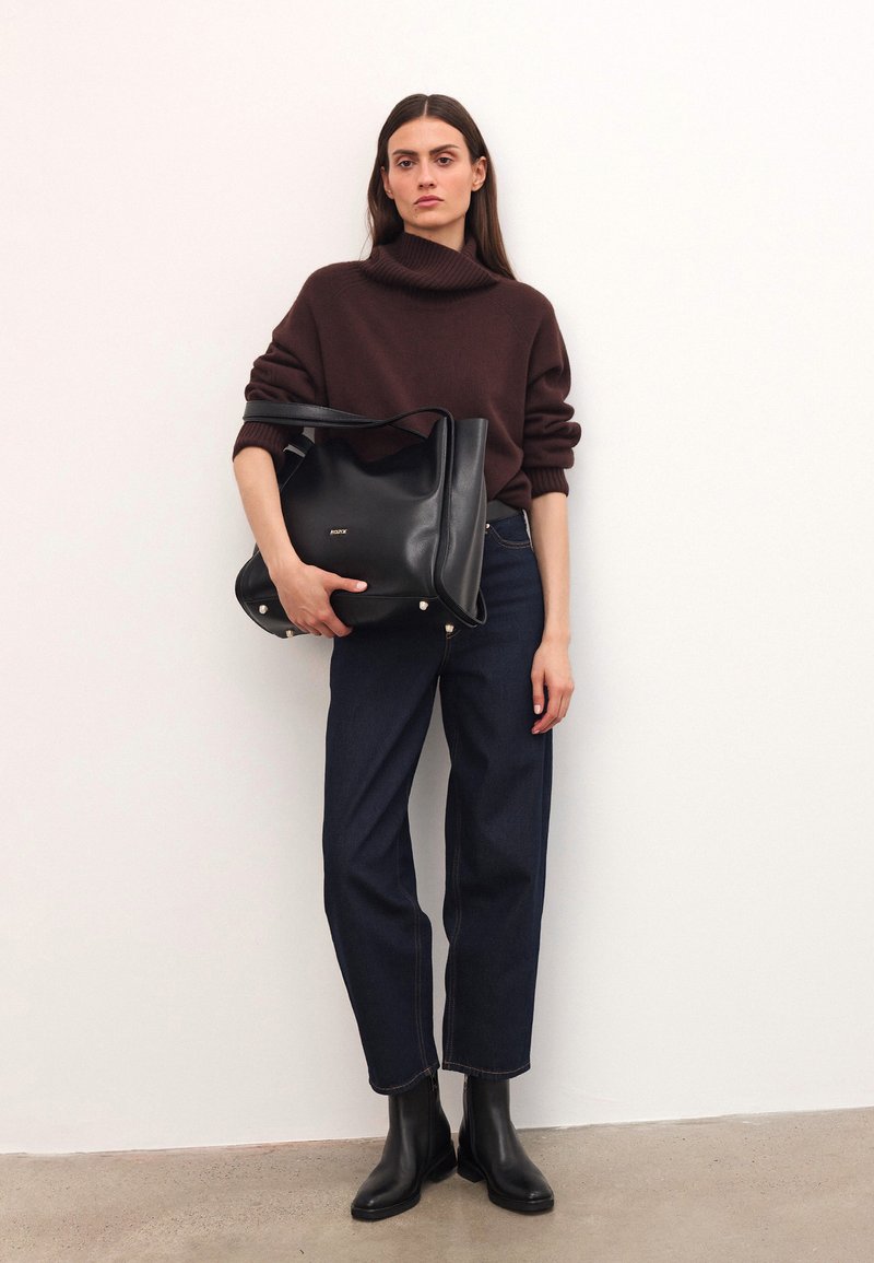 Naine, kes kannab pruunist kõrge kaelusega sviitrit, tumedaid siniseid teksaseid ja musti saapaid, hoiab suurt musta nahast käekotti valge seina vastas.