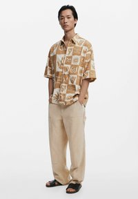 Kortærmet, mønstret skjorte i tan og hvid, med grafik; parret med lysebeige, løstsiddende bukser og sorte sandaler.