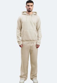 Beige Kapuzensweatshirt und passende Jogginghose aus weichem Stoff. Verfügt über eine Vordertasche und gerippte Bündchen. Getragen mit weißen Sneakers.