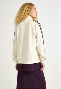 Krämfärgad sweatshirt med mörkbruna sidostrikar, mjuk textur, lös passform, hög krage, ribbade manschetter, i kombination med ett lager av ett mörk lila plagg.