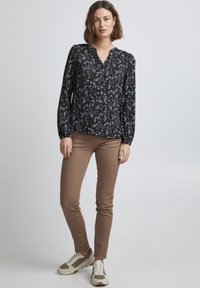 Blusa a maniche lunghe con motivo floreale scuro, caratterizzata da un scollo a V e pieghe. Abbinata a pantaloni beige aderenti e sneakers grigie, creando un look casual.