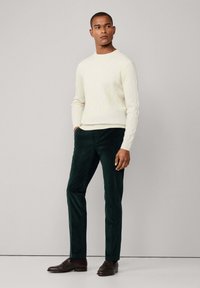 Muž stojaci v krémovom svetri s pleteninou, tmavozelených kordových nohaviciach a tmavohnedých kožených mokasínach na jednofarebnom pozadí.