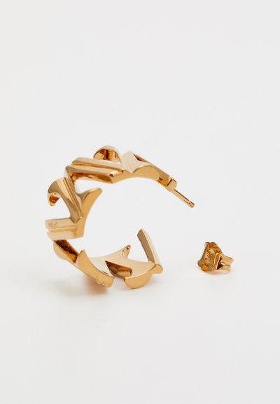 Boucles d'oreilles en métal doré en forme de créole avec un design ondulé ; elles possèdent un fermoir à clip et une petite puce d'oreille sur le côté.