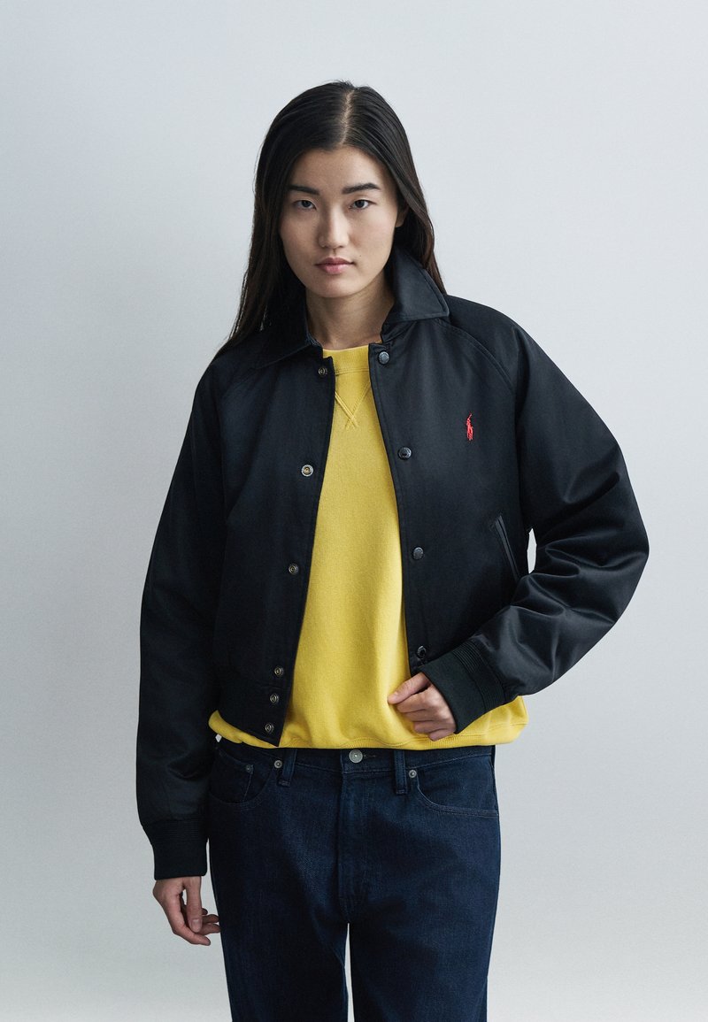 Čierna bomber bunda so zapínaním na patentky a rebrovanými manžetami, vrstvená cez žltú mikinu, v kombinácii s tmavomodrými džínsami. Vidite�ľný červený detail loga.