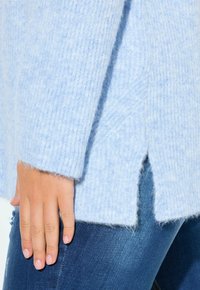 Hand auf dem Oberschenkel, trägt einen blauen Strickpullover mit Seitenschlitz und blaue Jeans.