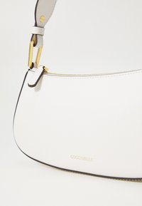 Borsa a spalla in pelle bianca con un design curvo, hardware tono oro e rifiniture nere. Presenta un logo del marchio visibile e dettagli con zip.