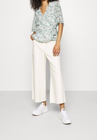 Top avvolgente floreale con maniche a sbuffo corte, abbinato a pantaloni bianchi a gamba larga e sneakers bianchi, creando un outfit leggero e informale.