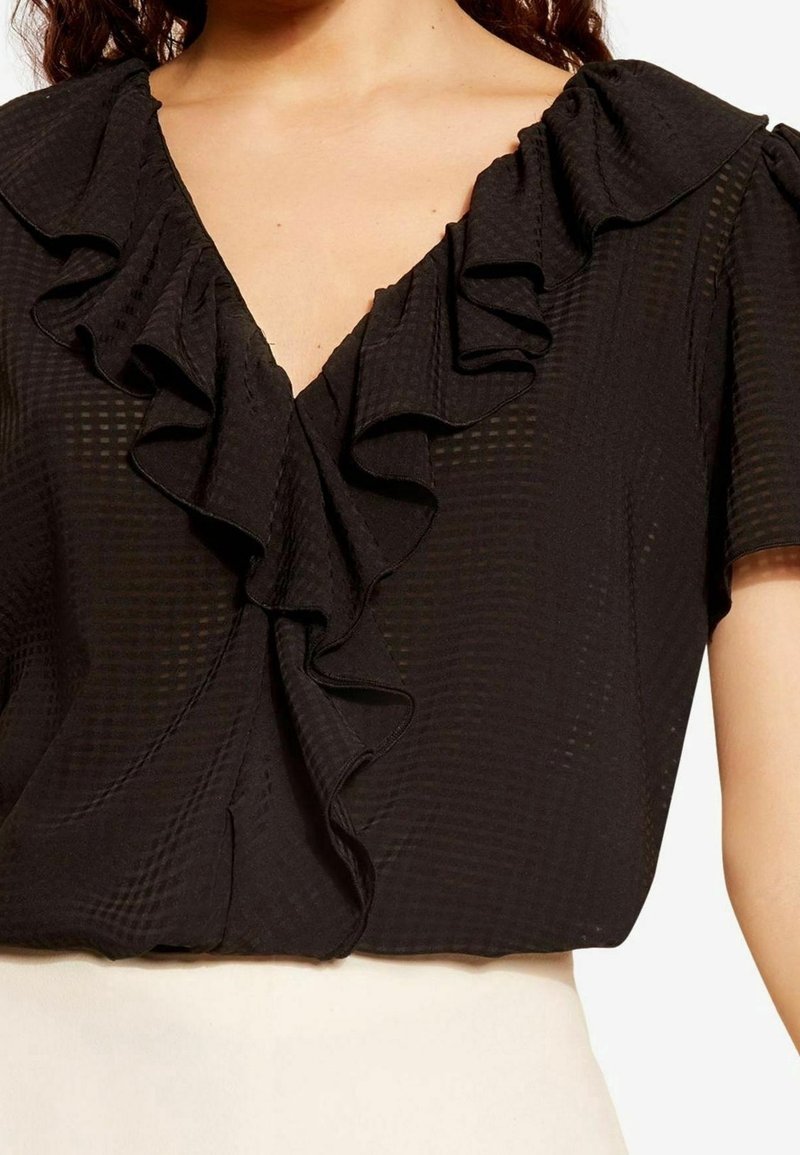 Blouse noire avec un col en V orné de volants, manches courtes bouffantes et tissu texturé à motif de grille subtil.