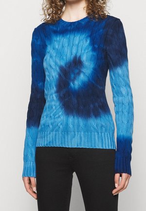 Frau trägt einen blauen Kabelstrickpullover mit dunklen und hellen blauen Batikmustern, kombiniert mit schwarzen Hosen vor einem weißen Hintergrund.