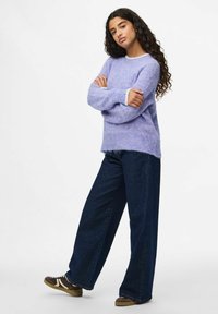 Femme jeune aux longs cheveux bouclés se tient les bras croisés, vêtue d'un pull en mohair lavande, de jeans larges foncés et de baskets marron.