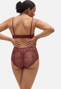 Un sujetador de encaje burdeos y bragas de cintura alta que presentan un patrón de hojas y correas ajustables con un cierre de gancho en la parte posterior.