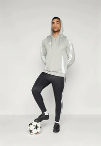 Szürke kapucnis pulóver fehér díszítéssel és oldalsó csíkkal, fekete nadrággal, amely fehér csíkokkal rendelkezik. Fekete focicipők, labda a lábaknál.