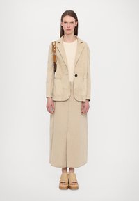 Beige kordflodjacka med två framfickor, som är tillsammans med en lång beige kjol. Träplattformsandaler med minimalistisk design och ljus färgpalett.