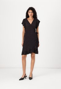 Vestido envolvente preto com estampa floral, mangas curtas com babados e uma barra com flounces, apresentando um detalhe de laço na cintura. Usado com sapatos de salto pretos tipo slingback.