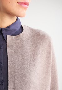 Beige svetr s měkkou texturou, otevřeným předním dílem a kulatým výstřihem, nošený přes námořnickou košili s límcem a knoflíky.