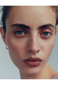 Kasvokuva vaaleaihoisesta henkilöstä, jolla on selkeät kulmakarvat, siniset silmät ja kiiltävät huulet. Hiukset ovat vedetty taakse. Hento meikki pehmeillä, luonnollisilla sävyillä.