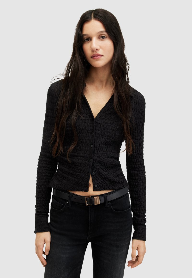 Cardigan lungo nero e testurizzato con scollo a V e bottoni frontali, abbinato a jeans scuri e una cintura nera con fibbia metallica.
