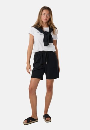 Ung kvinde iført en hvid t-shirt med print, sorte shorts med snøre, sort sweater bundet om skuldrene og sorte platformsandaler.