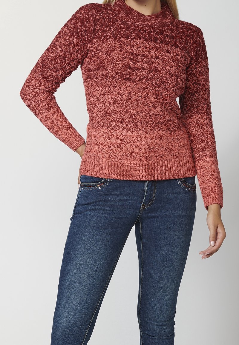 Ribbed knit sweater featuring a gradient ombre design in shades of red, with a textured pattern and a crew neckline. Paired with dark denim jeans.
