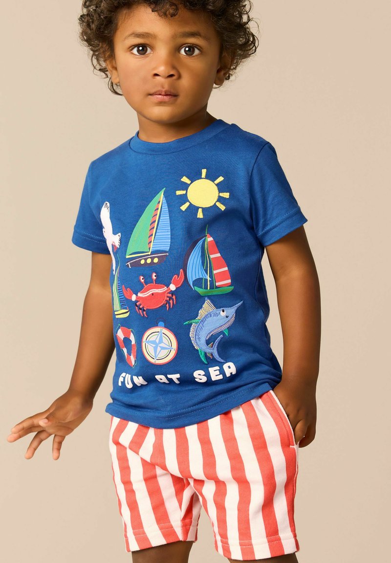 Jeune enfant portant un t-shirt bleu avec des illustrations sur le thème de la mer et le texte "Fun at Sea", ainsi qu'un short rayé rouge et blanc sur un fond neutre.