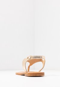 Kultainen metallinen thong-sandaali, jossa on pyöreä solki, tasapohja ja sileä pinta. Takaview, jossa on minimalistinen muotoilu ja luonnollisia väritampereita.