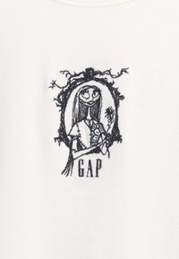 Hvitt stoff med et svart brodert motiv av en figur innenfor en dekorativ ramme, under hvilket teksten "GAP." står.