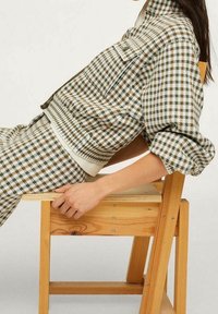 Skrzypcowy sweter w odcieniach ziemi z luźnym krojem, dużym kołnierzem i ściągaczami na rękawach, noszony podczas siedzenia na drewnianym krześle.