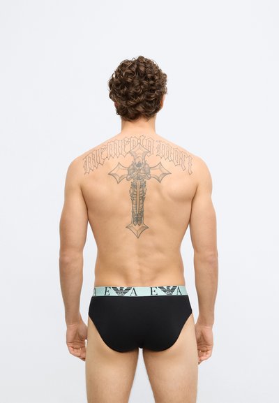 Cuecas pretas com cós azul claro apresentando um logótipo. Cabelo encaracolado e uma tatuagem detalhada nas costas são visíveis. Textura lisa.