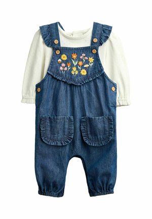 Wit overhemd met lange mouwen onder blauwe spijkeroveralls met gerimpelde bandjes, twee voorzakken, houten knopen en kleurrijke bloemenborduursels.