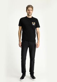 Camiseta negra con un estampado gráfico en el lado izquierdo del pecho, combinada con pantalones negros y zapatillas negras. Diseño simple y forma ajustada.