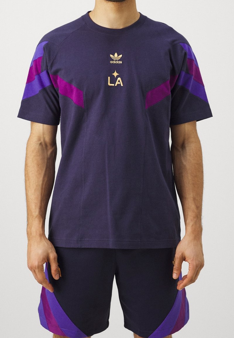 Marinblå sporttröja med lila detaljer, med rund halsringning, korta ärmar och "LA"-logotyp i guld. Matchas med matchande shorts.