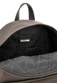 Zaino in pelle marrone con una texture liscia, dotato di chiusura con zip e un interno nero a motivo etichettato "GUESS ECO."