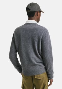 Grijze gebreide trui met een ronde halslijn, geribbelde mouw- en tailleboorden. Draagt een witte blouse eronder, met groenbruin gekleurde broek.