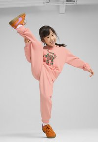 Sudadera rosa y pantalones deportivos con un patrón de elefantes verdes y rojos; zapatillas naranjas con acentos morados; niña realizando un levantamiento de pierna alto.