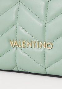 Tűzött menta zöld bőr táska, amelyen arany "VALENTINO" felirat található. A dizájn jellemvonása a cikkcakk varrás és a sima, fényes felület.