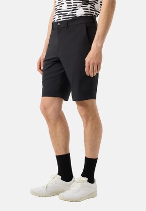 Mann trägt schwarze knielange Shorts, schwarze Socken, weiße Sneakers und ein schwarz-weiß gemustertes Kurzarmhemd und steht vor einem einfarbigen Hintergrund.
