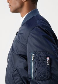 Marine bomberjakke med en jevn, blank tekstur. Har blå krage, glidelås-lommepose på armen og patch-logo på ermet.