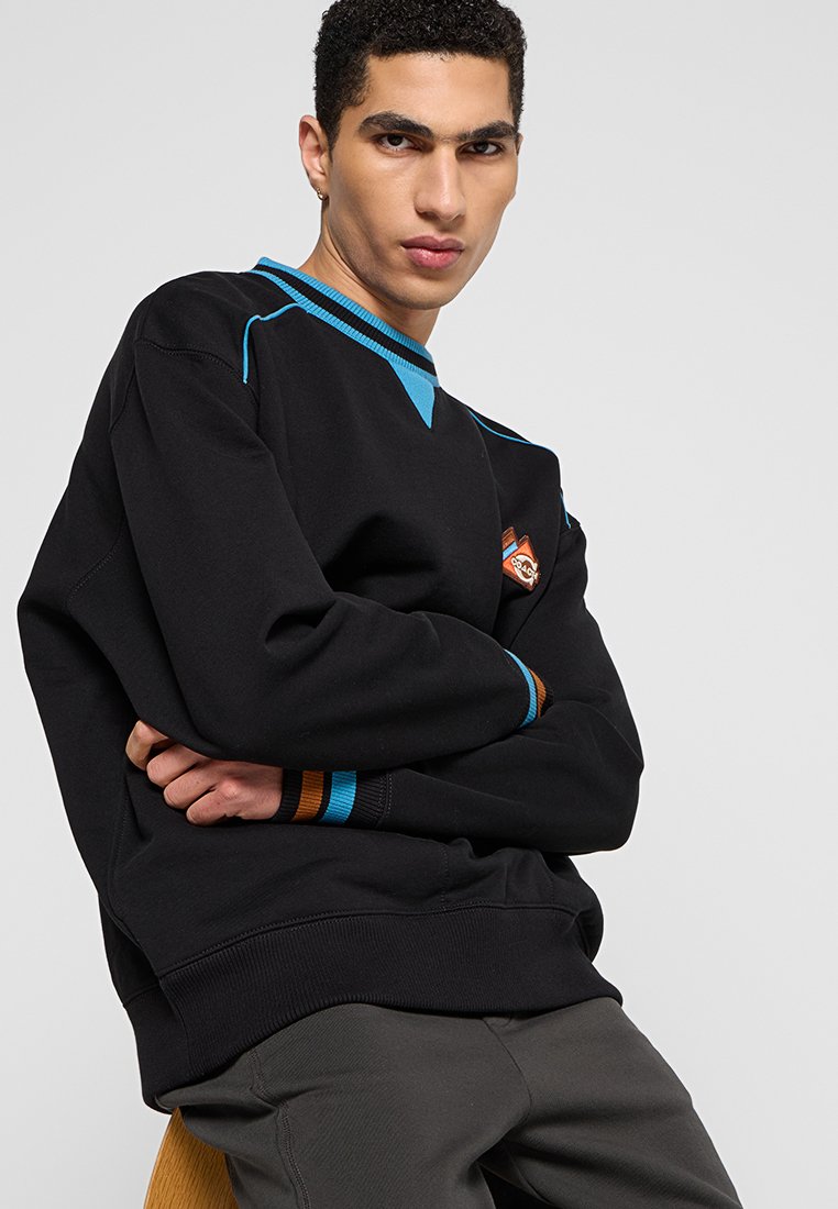 Czarna bluza z niebieskimi akcentami i diamentowym pomarańczowym logo. Posiada ściągacze przy mankietach i dekolcie, miękką tkaninę oraz luźny krój.