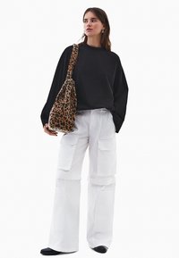 Czarna koszula z długim rękawem, białe szerokie spodnie cargo z kieszeniami, torba typu tote w leopardzim wzorze. Model stoi w czarnym obuwiu na białym tle.