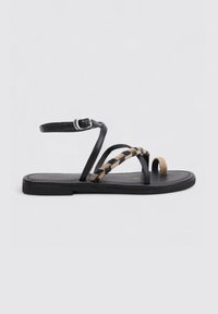 Sandalias de cuero negro con correas trenzadas en beige y negro. Cuentan con una hebilla ajustable en el tobillo y una suela plana de goma. Diseño de punta abierta.