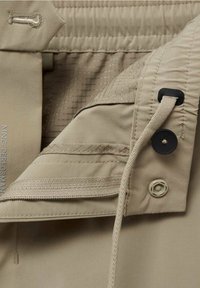 Détail de tissu beige montrant une ceinture avec bouton, boutonnière, cordon élastique, bouton-pression et doublure intérieure texturée.
