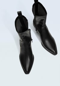 Botas de cuero negro hasta el tobillo con punta afilada, con cremalleras laterales y una textura suave. Con detalles de costura sutil.