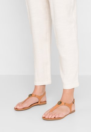 Voeten met beige platte sandalen met decoratieve ronde metalen accenten, gecombineerd met losse witte broeken die boven de enkels zijn afgeknipt.