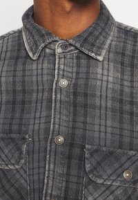 Graues kariertes Flanellhemd mit Button-Down-Kragen, Metallknöpfen und zwei Brusttaschen, zeichnet sich durch eine weiche Textur und kontrastierende Nähte aus.