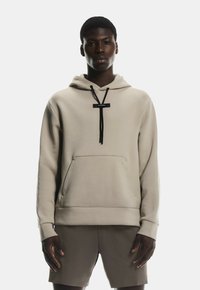Sweat à capuche beige avec une poche avant, des cordons de serrage noirs et un design minimaliste. Porté avec un short beige assorti.