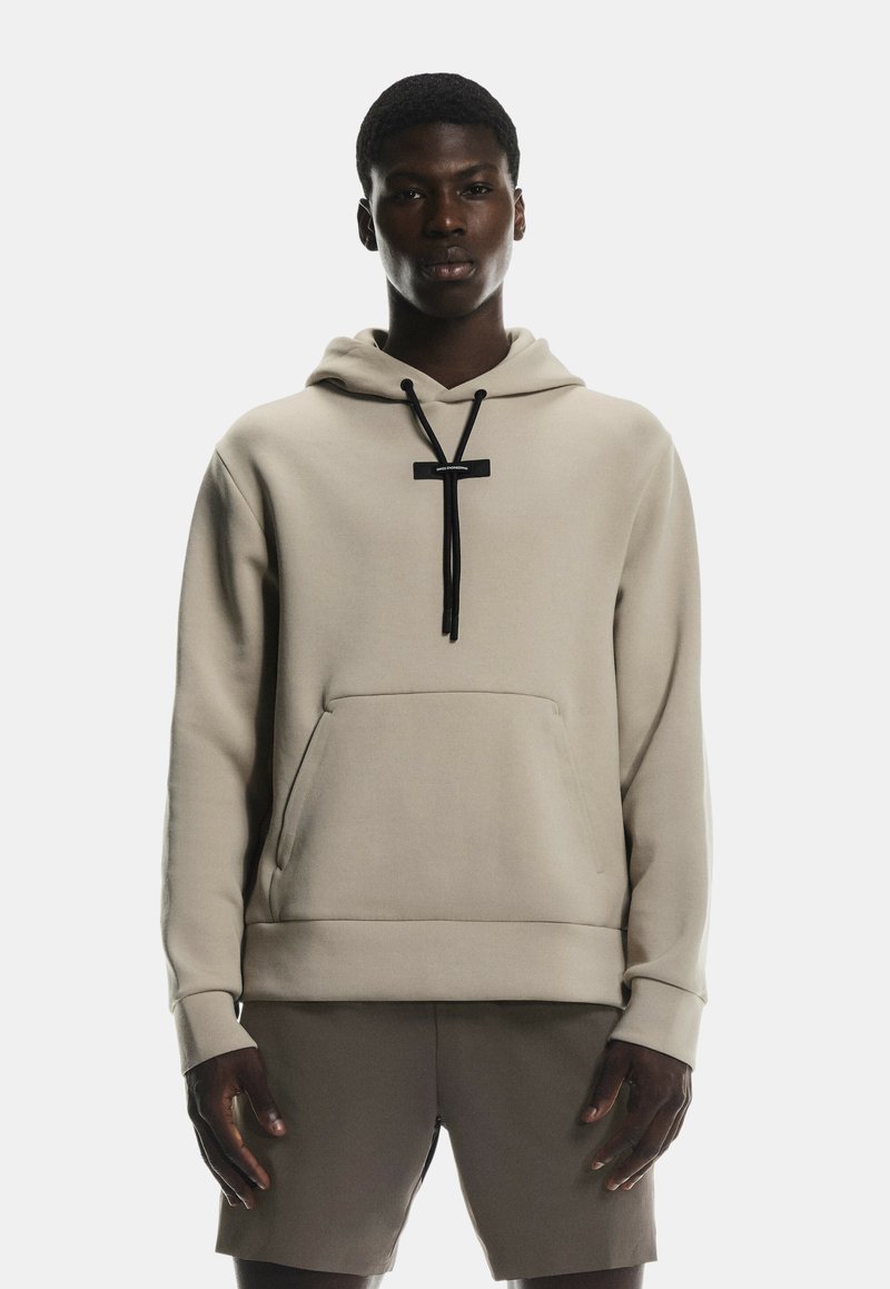 Sweat à capuche beige avec une poche avant, des cordons de serrage noirs et un design minimaliste. Porté avec un short beige assorti.